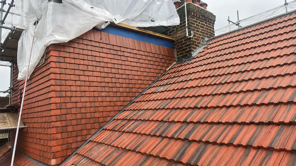 Red clay tile-hung dormer and red interlocking roof tiles beside a brick chimney, with scaffold sheeting above