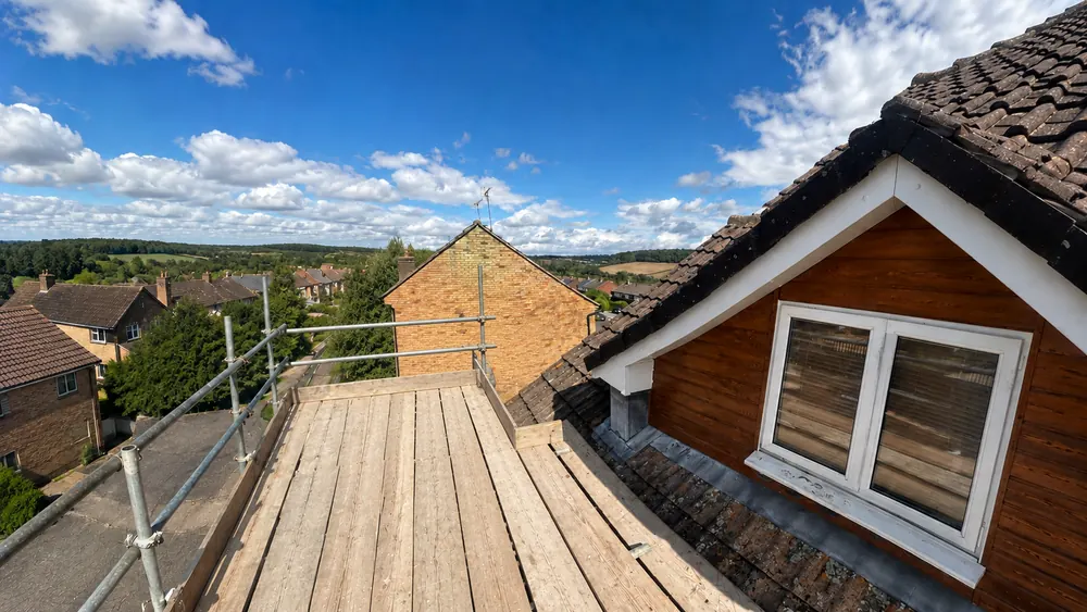 Timber-clad dormer with lead flashing beside a scaffold platform, rooftops and trees beyond